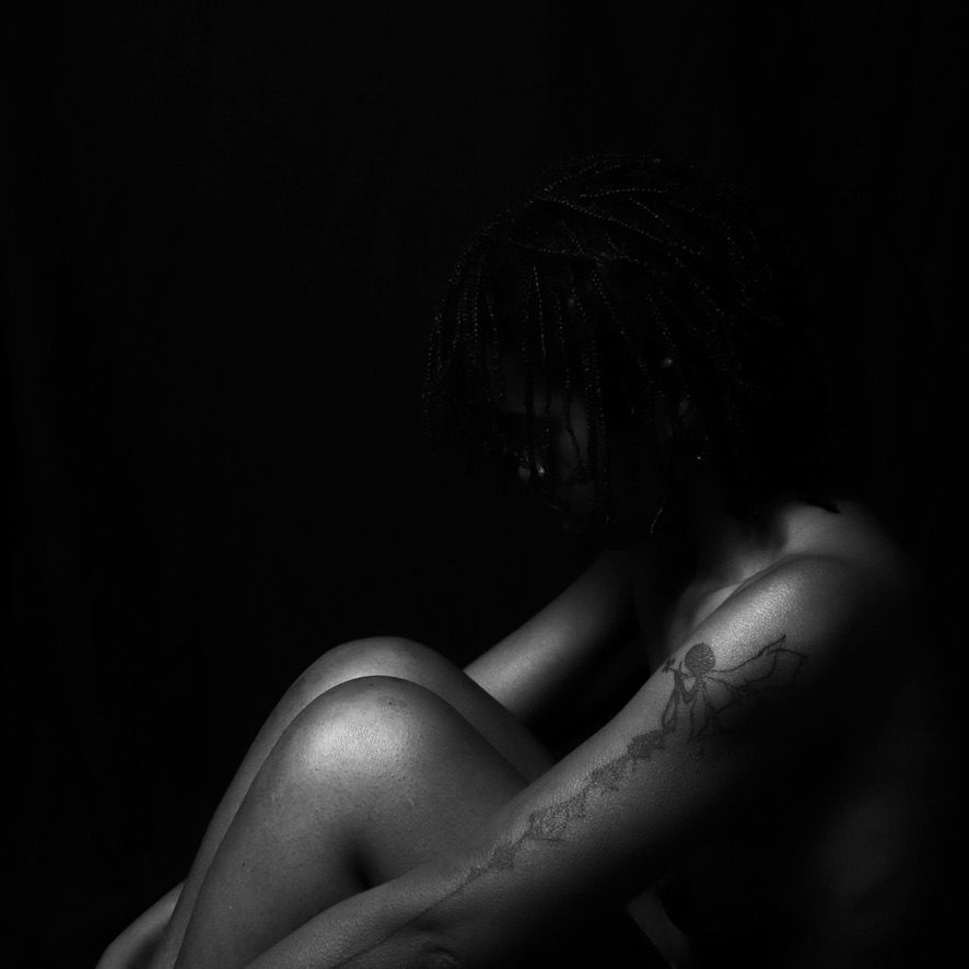 Pessoa sentada com os joelhos recolhidos, o rosto oculto pelas tranças e uma tatuagem visível no braço, numa fotografia a preto e branco.