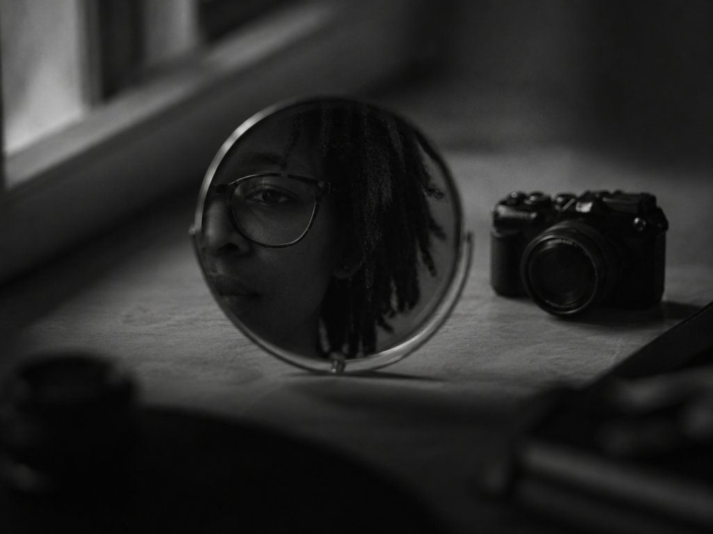 Rosto refletido num espelho redondo, com uma câmara fotográfica pousada ao lado, numa fotografia a preto e branco.